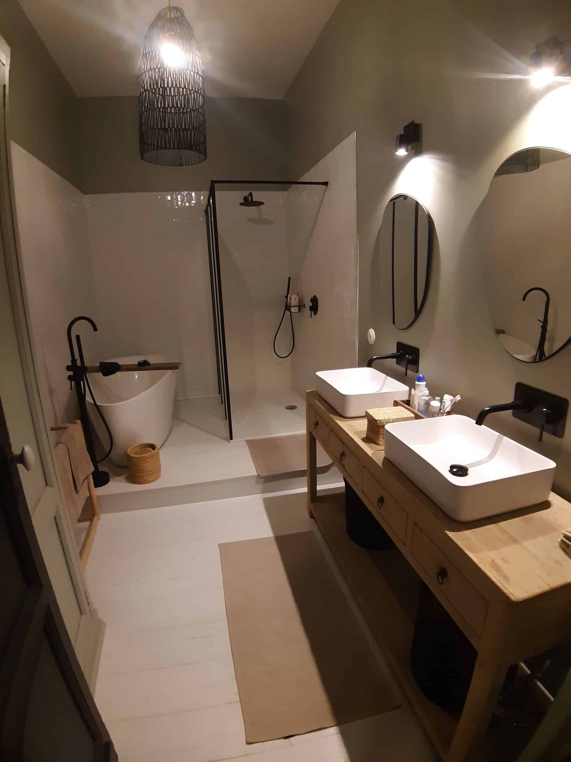 Salle de bain moderne avec baignoire îlot, douche vitrée noire, et double vasque sur meuble en bois clair, éclairage chaleureux.