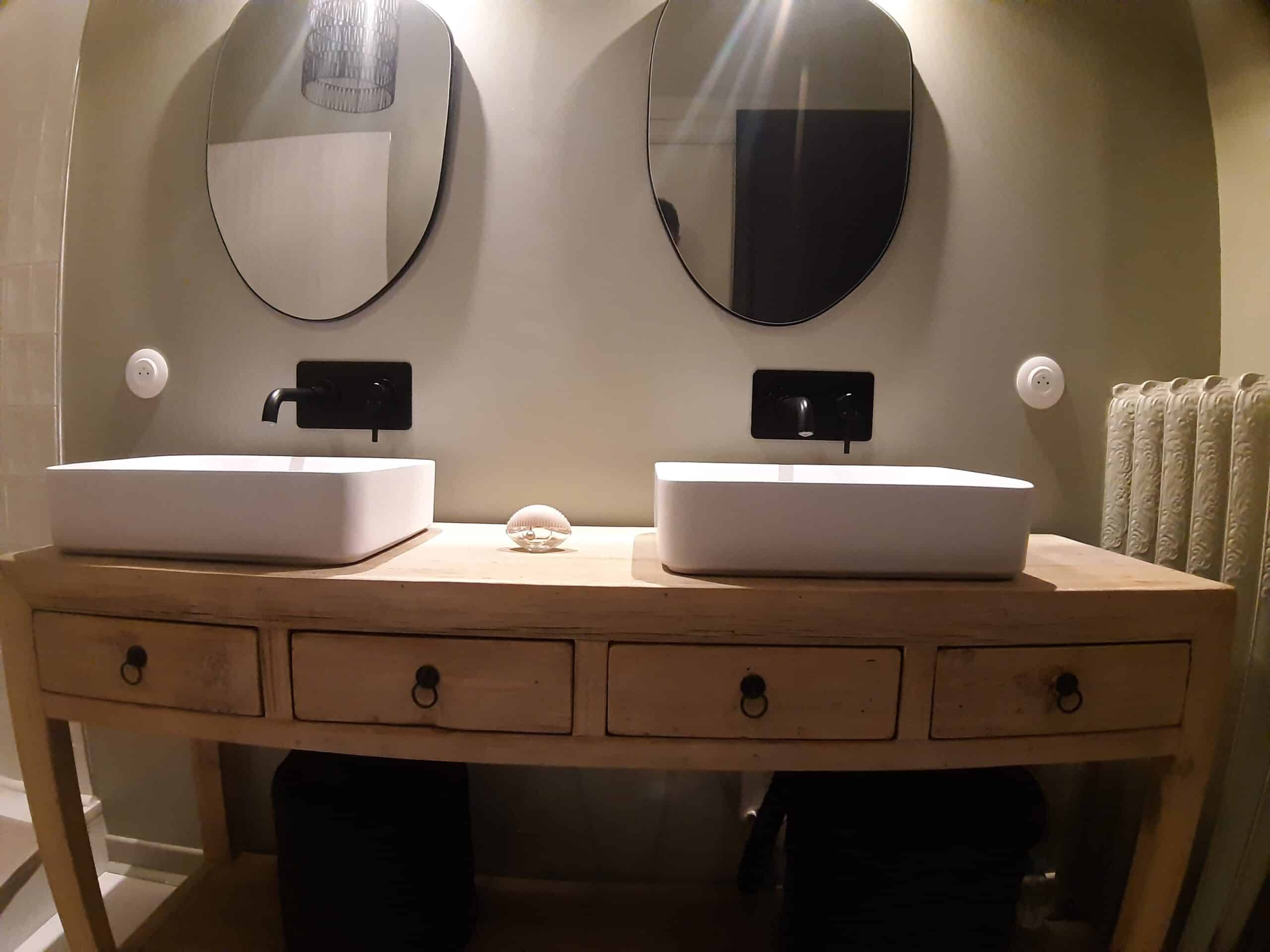 Salle de bain avec double vasque rectangulaire blanche sur console en bois patiné, robinetterie noire murale et miroirs ovales.
