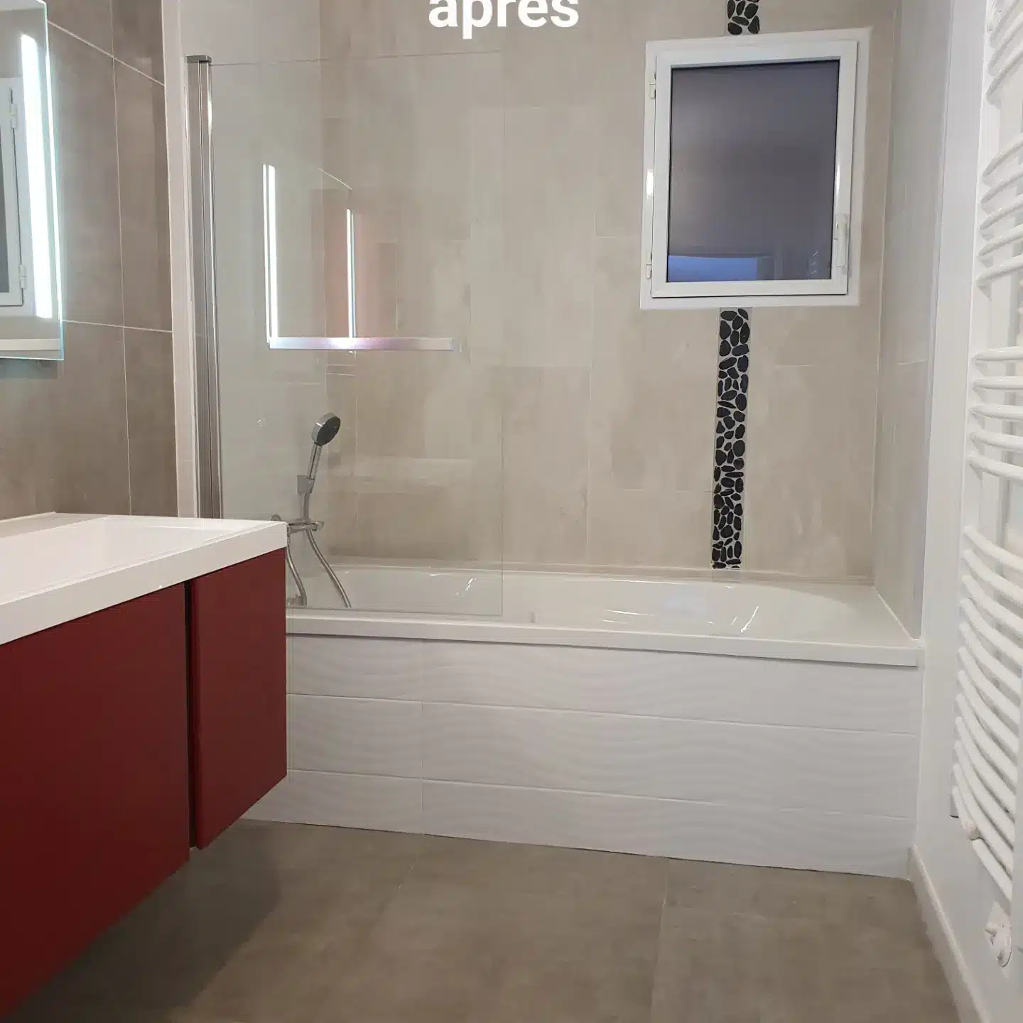 Salle de bain rénovée après travaux : baignoire, paroi vitrée, meuble vasque rouge bordeaux, carrelage beige et mosaïque galets noirs.