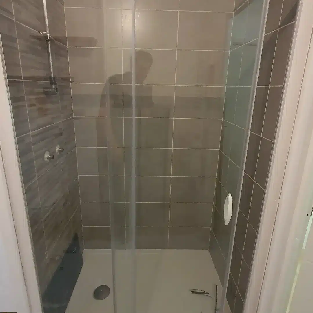 Vue d'une douche moderne avec parois vitrées et carrelage mural gris clair et taupe. Reflet d'une personne visible sur la vitre.