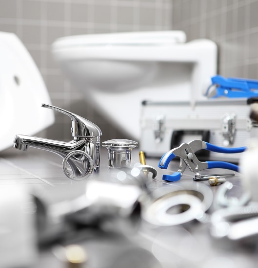 Outillage de plombier, mitigeur chromé et pièces détachées devant des toilettes et un lavabo flous en arrière-plan.