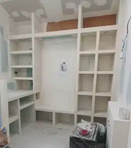 Bibliothèque encastrée en placo et plâtre, brute et non peinte, avec étagères de différentes tailles et espace central, chantier en cours. Une feuille est épinglée au centre.