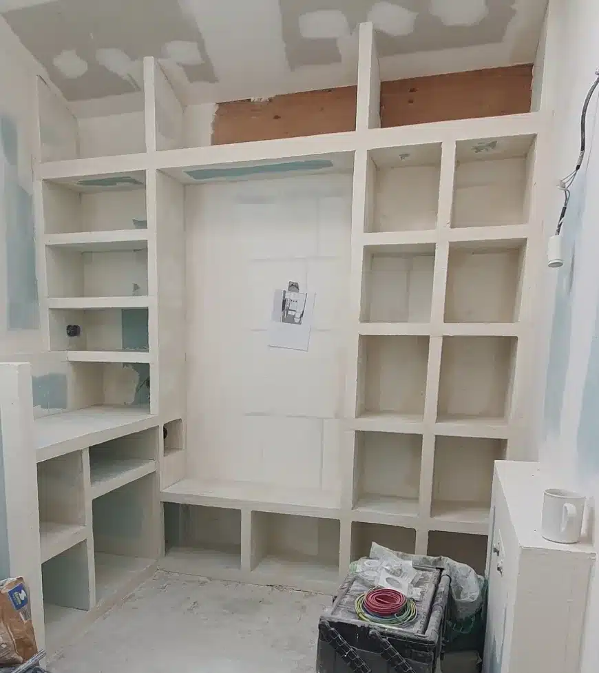 Bibliothèque encastrée en placo et plâtre, brute et non peinte, avec étagères de différentes tailles et espace central, chantier en cours. Une feuille est épinglée au centre.