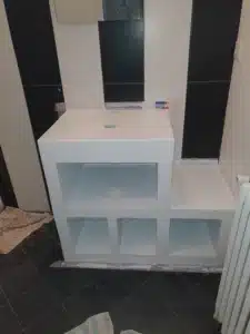Meuble vasque blanc sur mesure avec étagères ouvertes et trou pour robinetterie, dans une salle de bain carrelage noir et murs rayés blanc/noir.