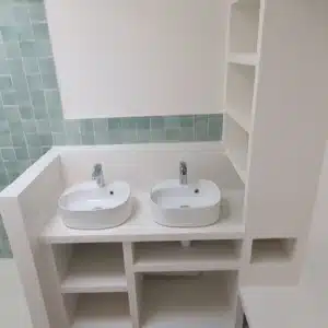 Salle de bain moderne avec une longue vanité blanche, deux vasques ovales posées, robinetterie chromée, et étagères intégrées, accentuée par un carrelage mural bleu-vert aqua.