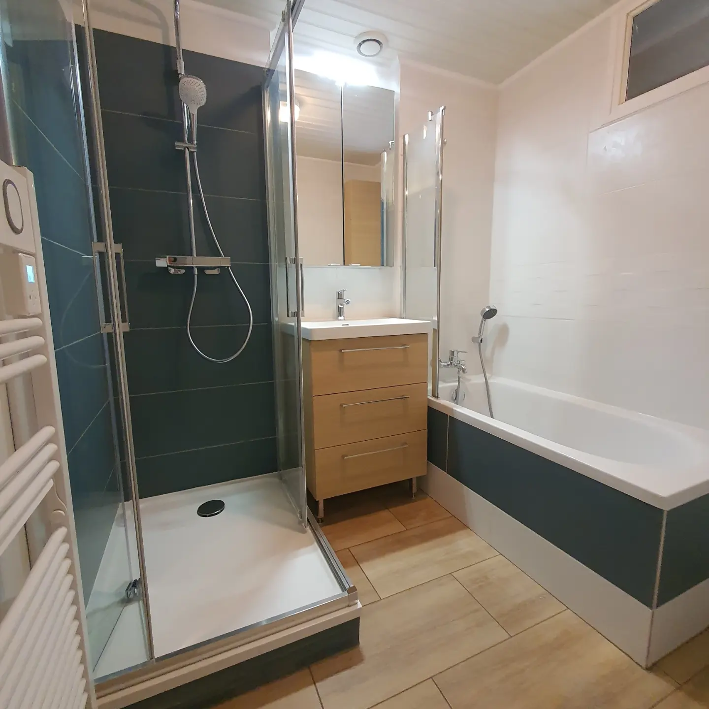 Salle de bain moderne avec douche à l'italienne vitrée, meuble vasque bois clair et baignoire au tablier gris foncé, sol carrelé beige.