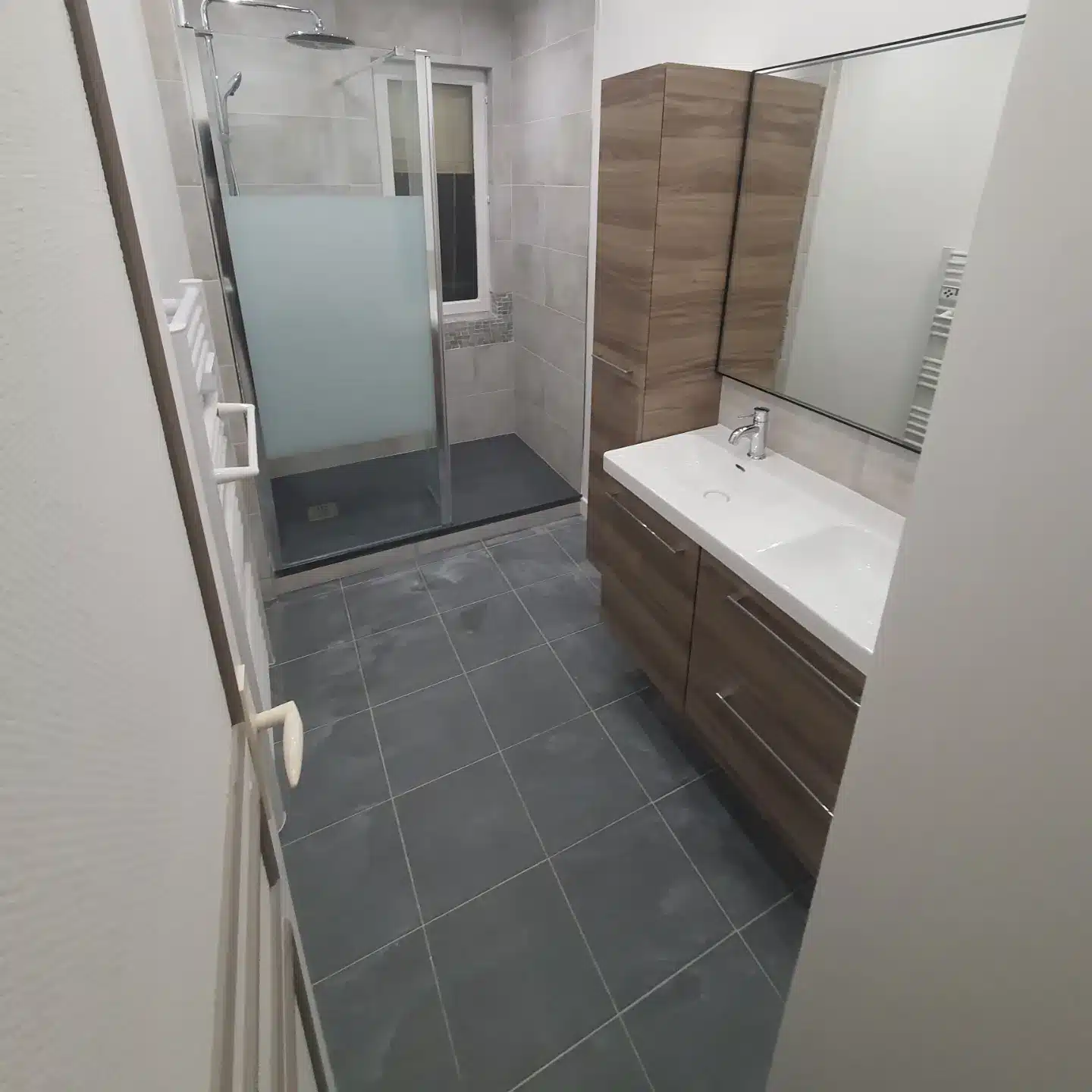 Salle de bain moderne avec douche vitrée à paroi dépolie, meuble-lavabo en bois, miroir horizontal et carrelage gris foncé au sol.